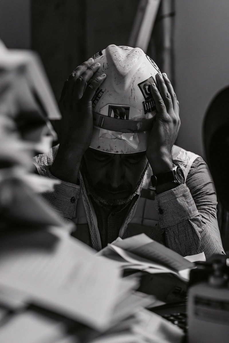 An overwhelmed tradie at his desk, head in hands, buried in paperwork